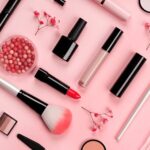 Various cosmetic accessories for makeup and manicure on trendy pastel pink background with red flowers. Blush, brush, eye shadow, mascara, perfume, lipstick, nail Polish. Skin care products.